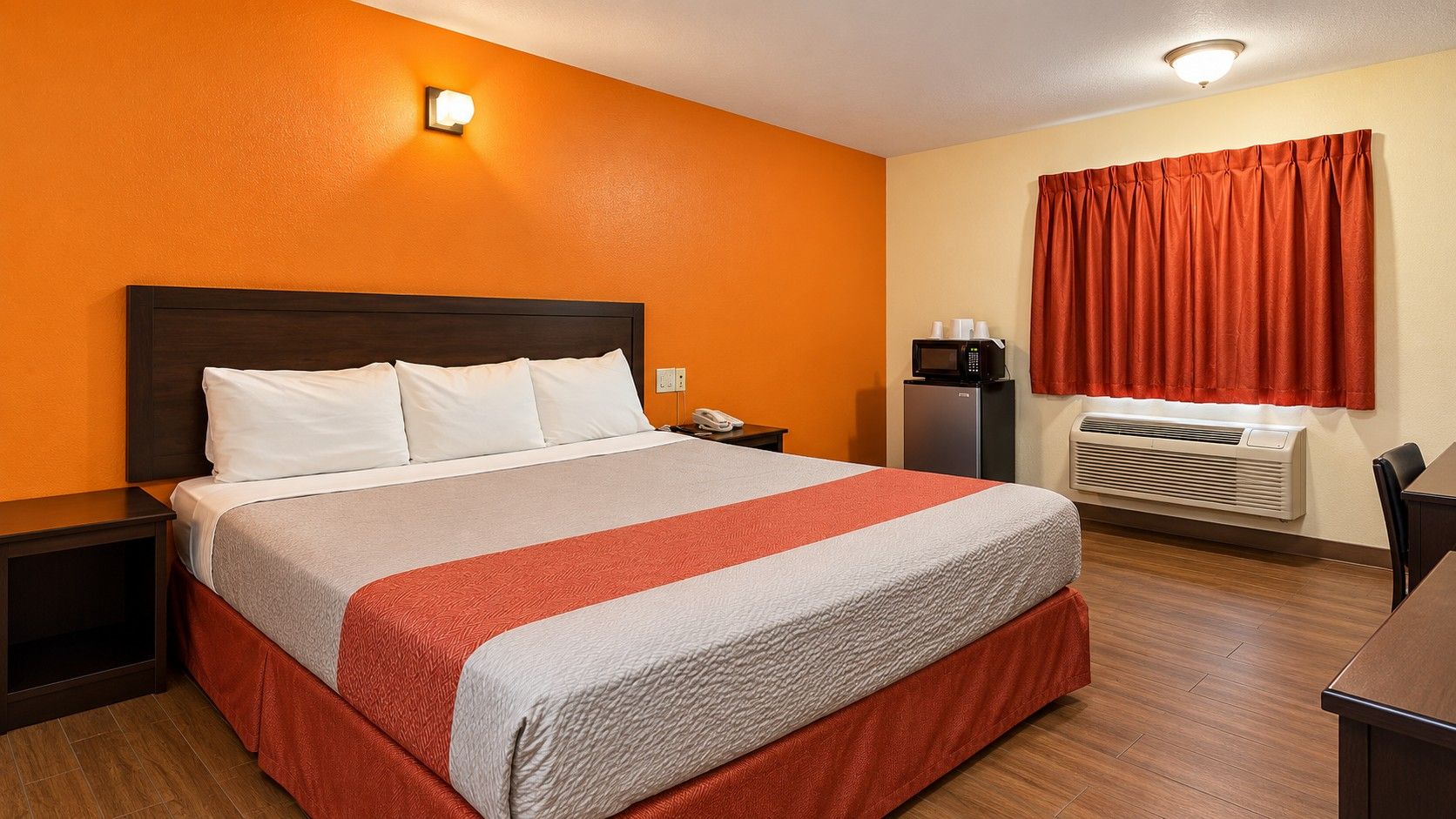 A hotel room with a bed, nightstand, bedside table, microwave, and an air conditioning unit.