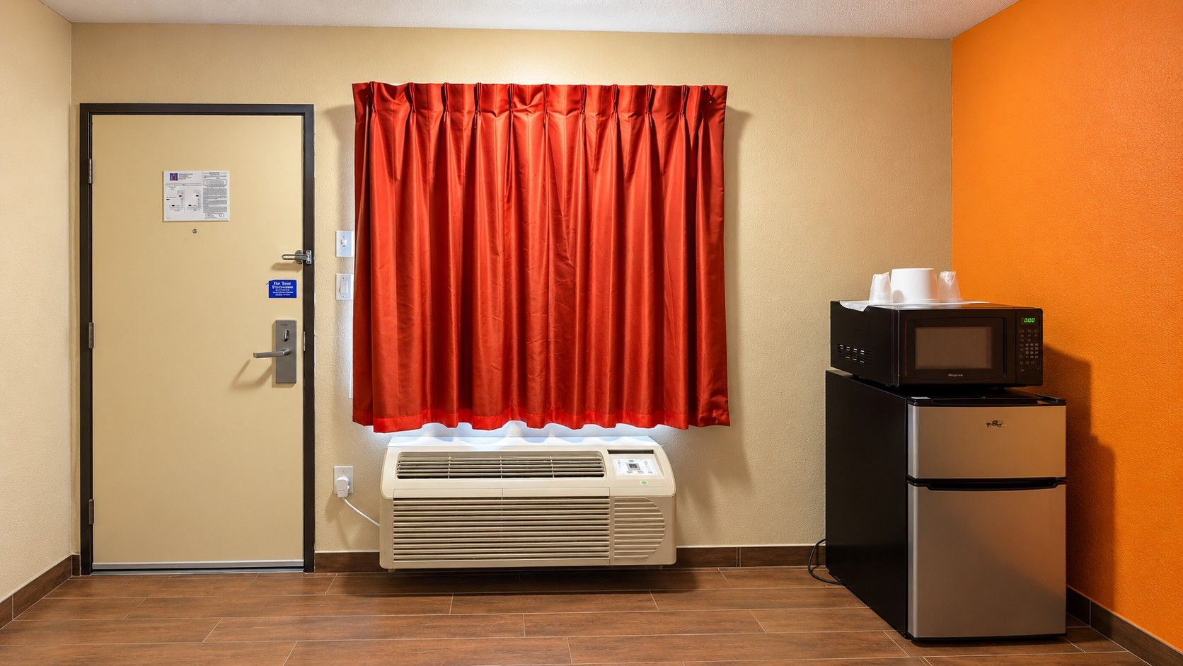 Une chambre d’hôtel équipée d’une porte, d’un climatiseur, d’un réfrigérateur et d’un four à micro-ondes posé sur un plan de travail.