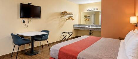 A hotel room with a bed, a desk with chairs, a television, a vanity with a mirror, and a wall-mounted light.