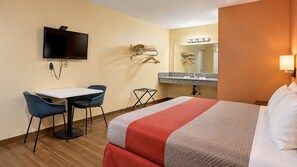 A hotel room with a bed, a desk with chairs, a television, a vanity with a mirror, and a wall-mounted light.