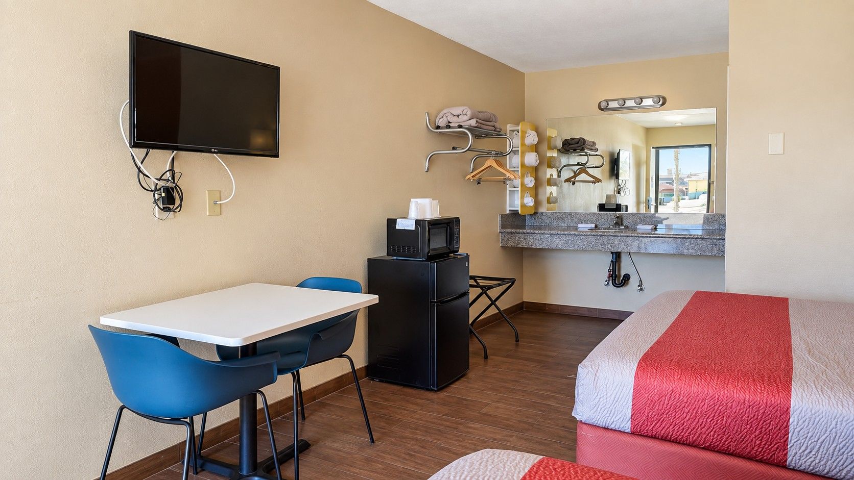 Habitación de hotel con cama, escritorio con silla, refrigerador, microondas, televisor montado en la pared y baño con espejo y toallero.