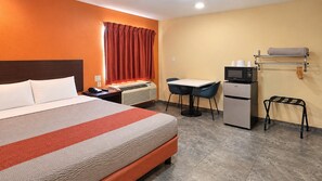 A hotel room with a bed, desk, chair, microwave, refrigerator, and a small table with a towel.