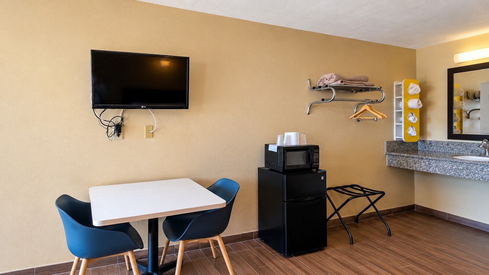 A compact hotel room with a flat-screen TV, a small kitchenette with a microwave, a dining area with a table and chairs, and a bathroom with a sink and mirror.