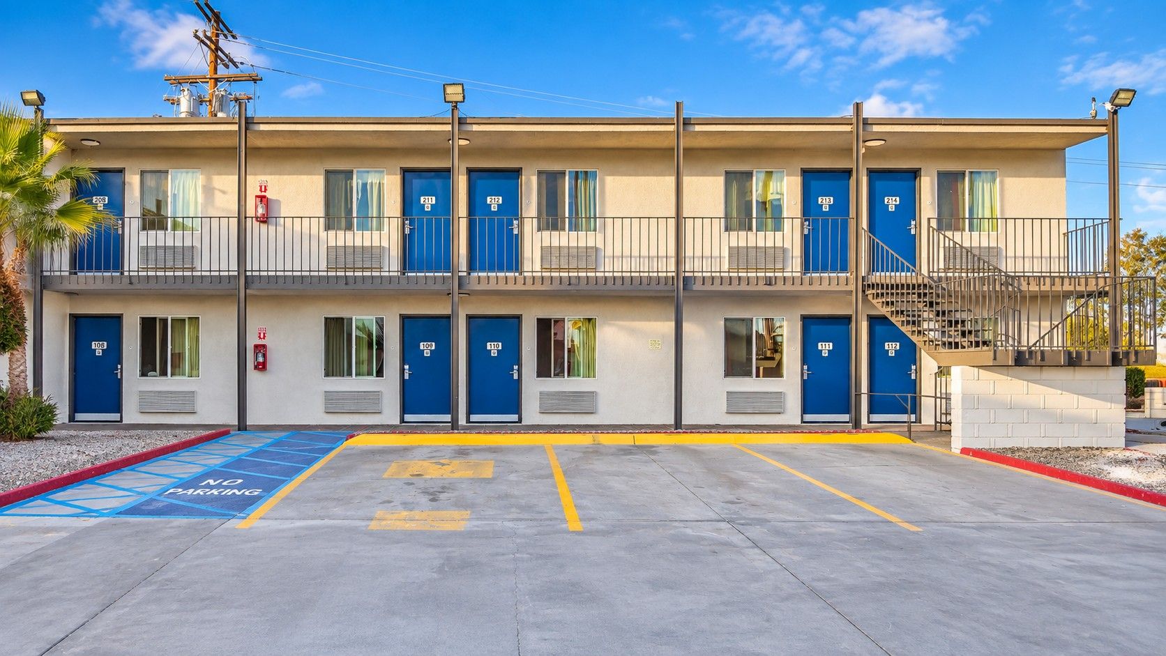 Un motel con varias puertas azules, un estacionamiento sin señales de prohibido aparcar, y un cielo despejado.