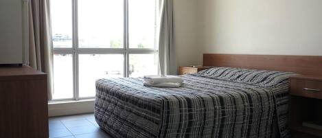 A hotel room with a bed, a wooden headboard, a nightstand, a desk, and an air conditioning unit.