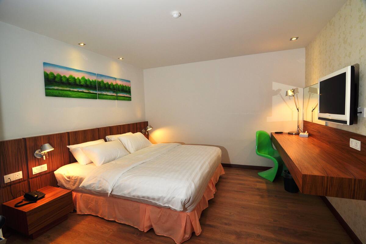 A hotel room with a wooden bed, a desk with a TV, a green chair, and a painting of a landscape on the wall.