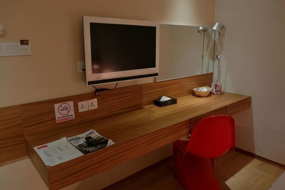 A hotel room with a wooden desk, a red chair, a flat-screen TV, a mirror, and a lamp.