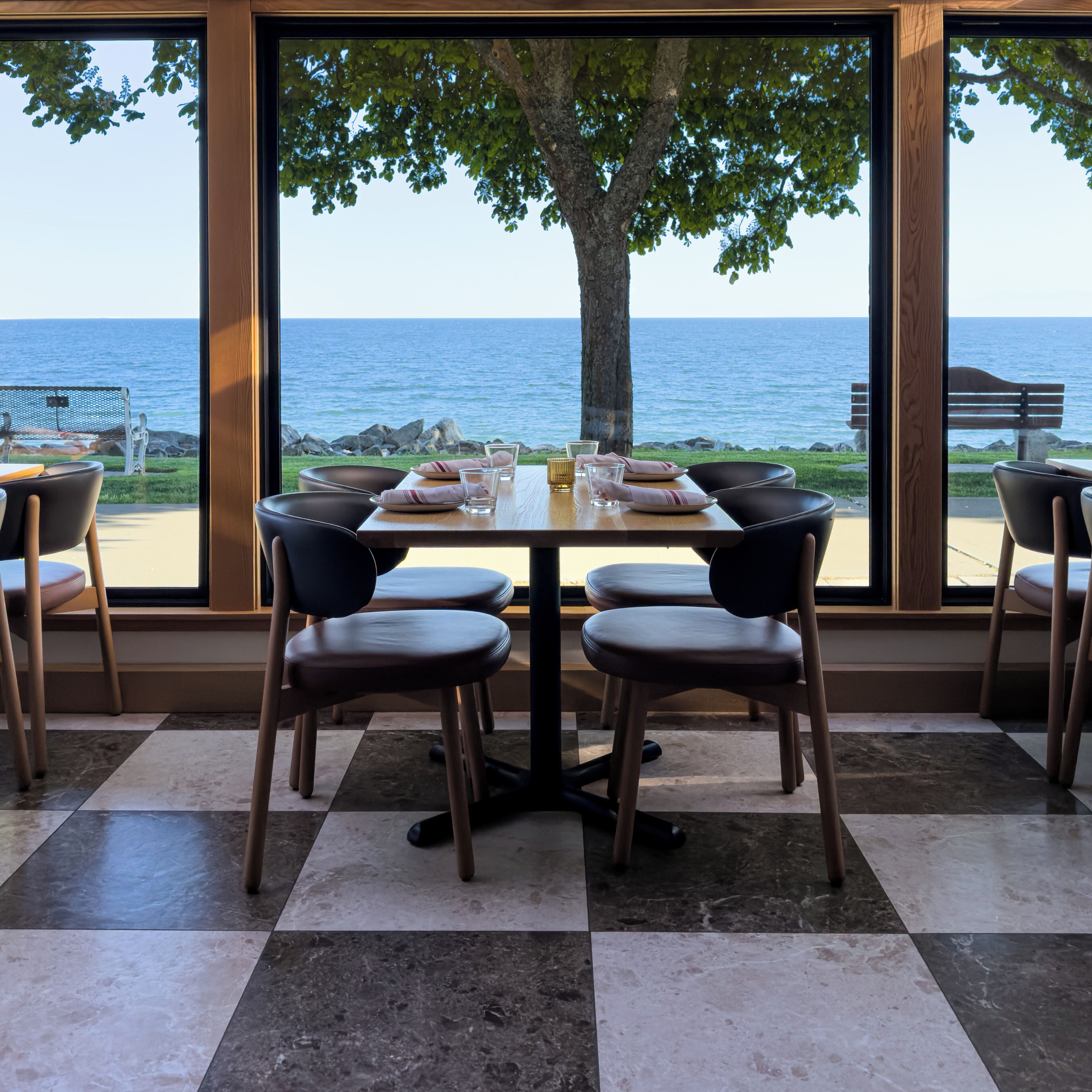 Un comedor con una mesa dispuesta para cuatro, ventanas amplias con vistas al mar y un árbol afuera.