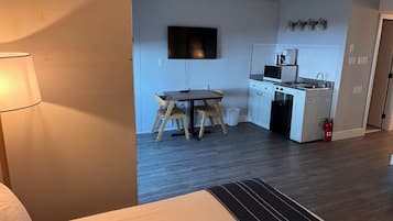 A compact hotel room with a bed, a small dining table, a TV, a kitchenette, and a fire extinguisher.