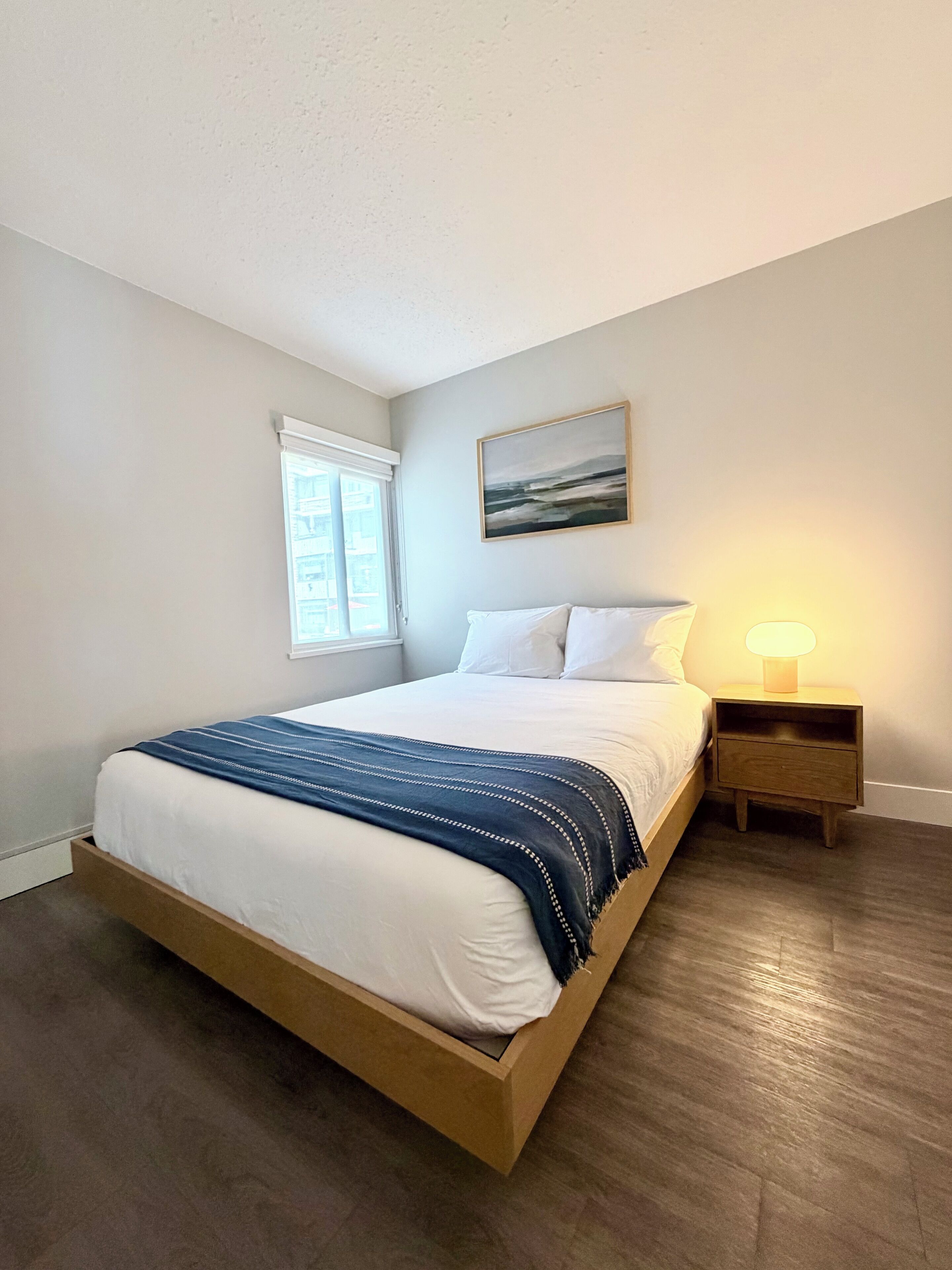 A bedroom with a bed, a nightstand, a lamp, a window, and a painting on the wall.
