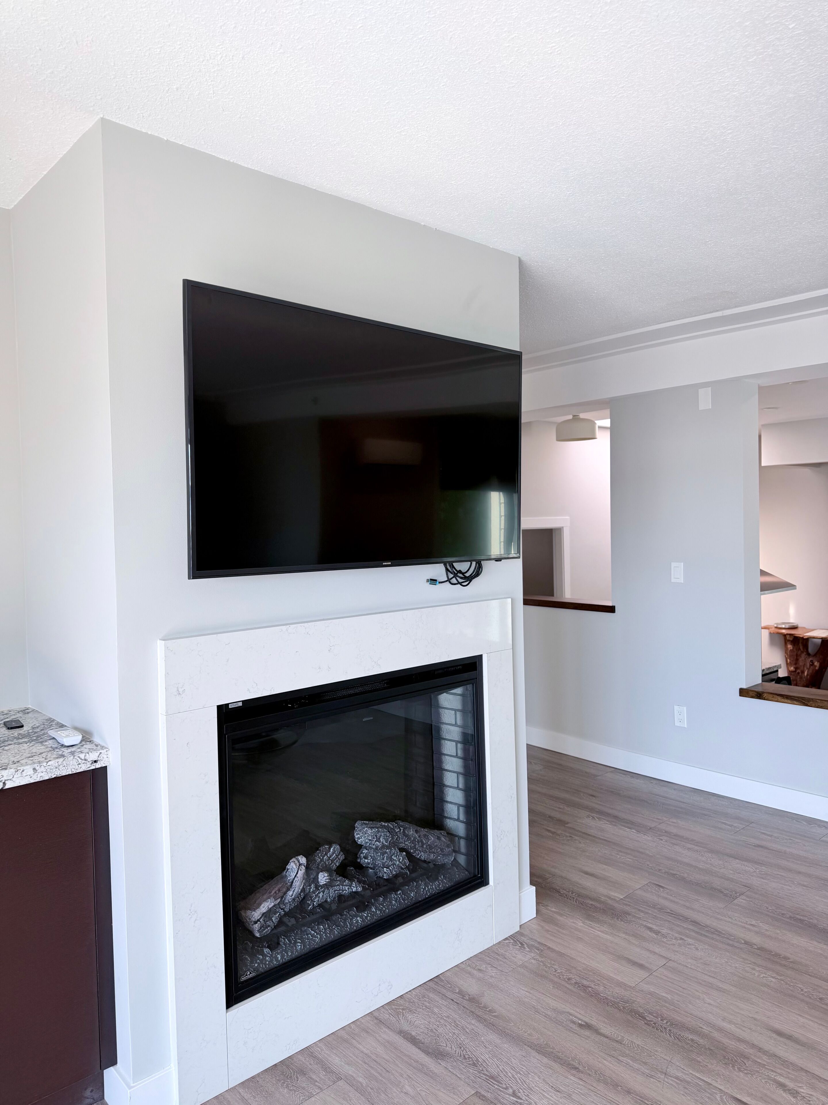 Una sala de estar con una televisión de pantalla plana sobre la chimenea, una cocina al fondo y piso de madera.