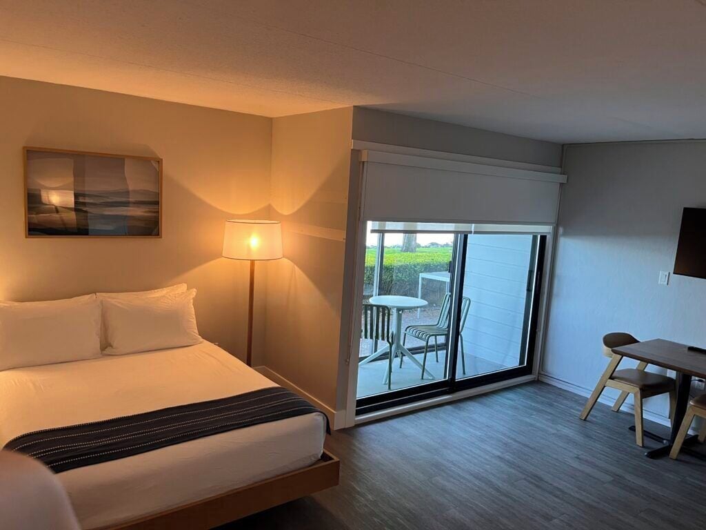 A hotel room with a bed, a desk, a chair, a lamp, and a view through a sliding glass door.