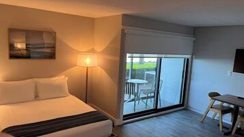 A hotel room with a bed, a desk, a chair, a lamp, and a view through a sliding glass door.