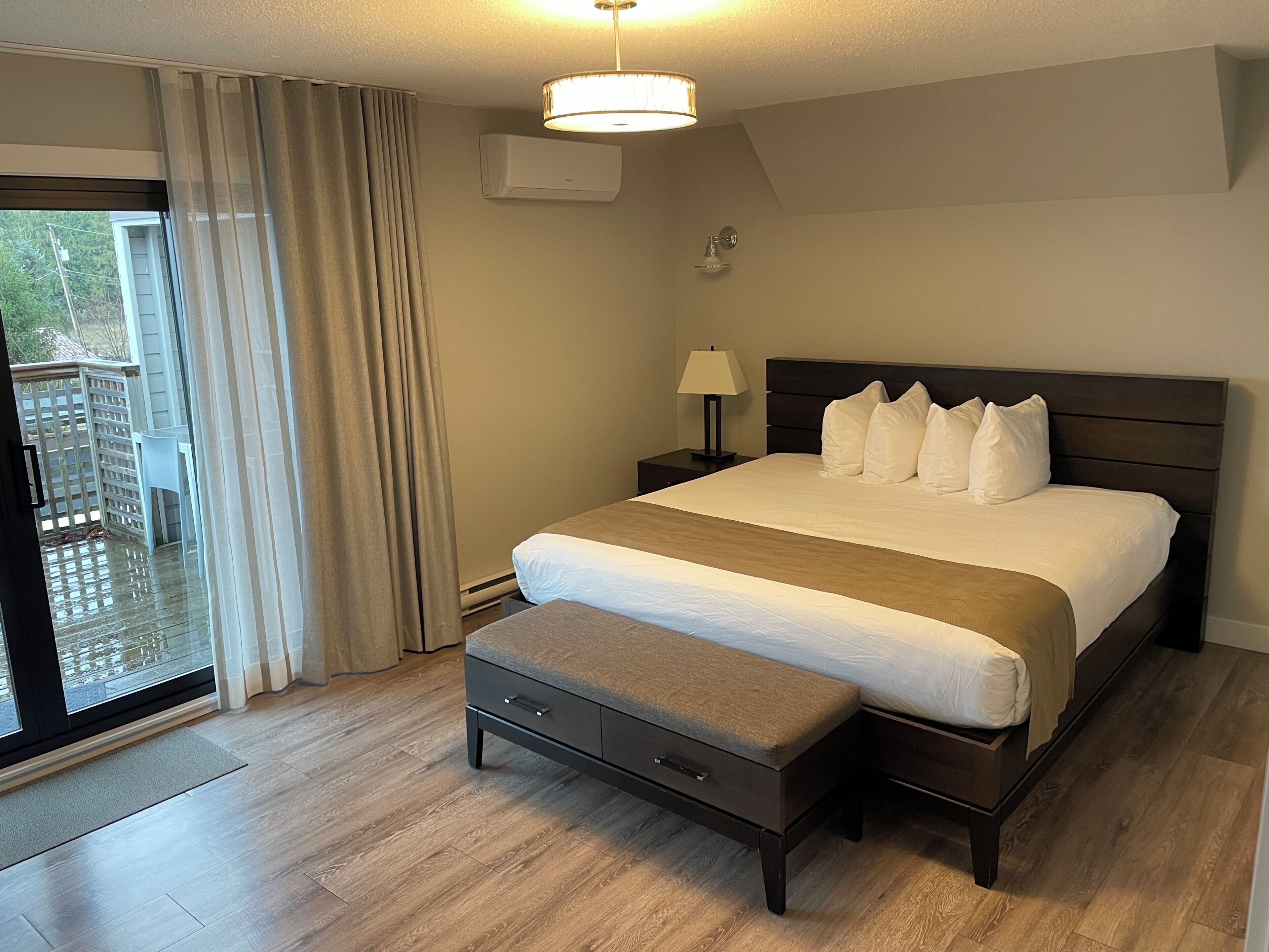 A hotel room with a bed, a nightstand, a lamp, a bench, and a sliding glass door to an outdoor area.