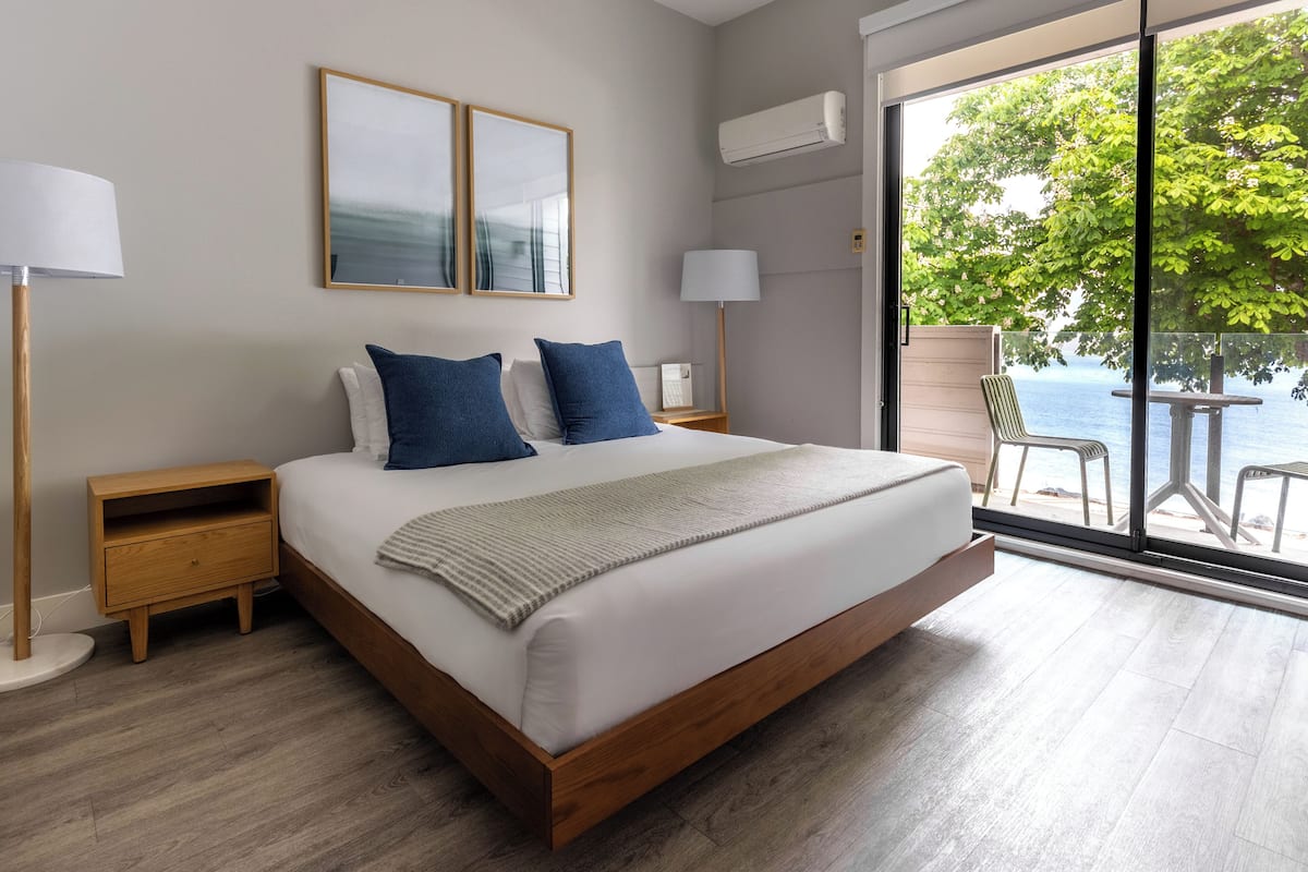 A modern bedroom with a large bed, two nightstands, a window with a view of the sea, and a balcony with outdoor seating.