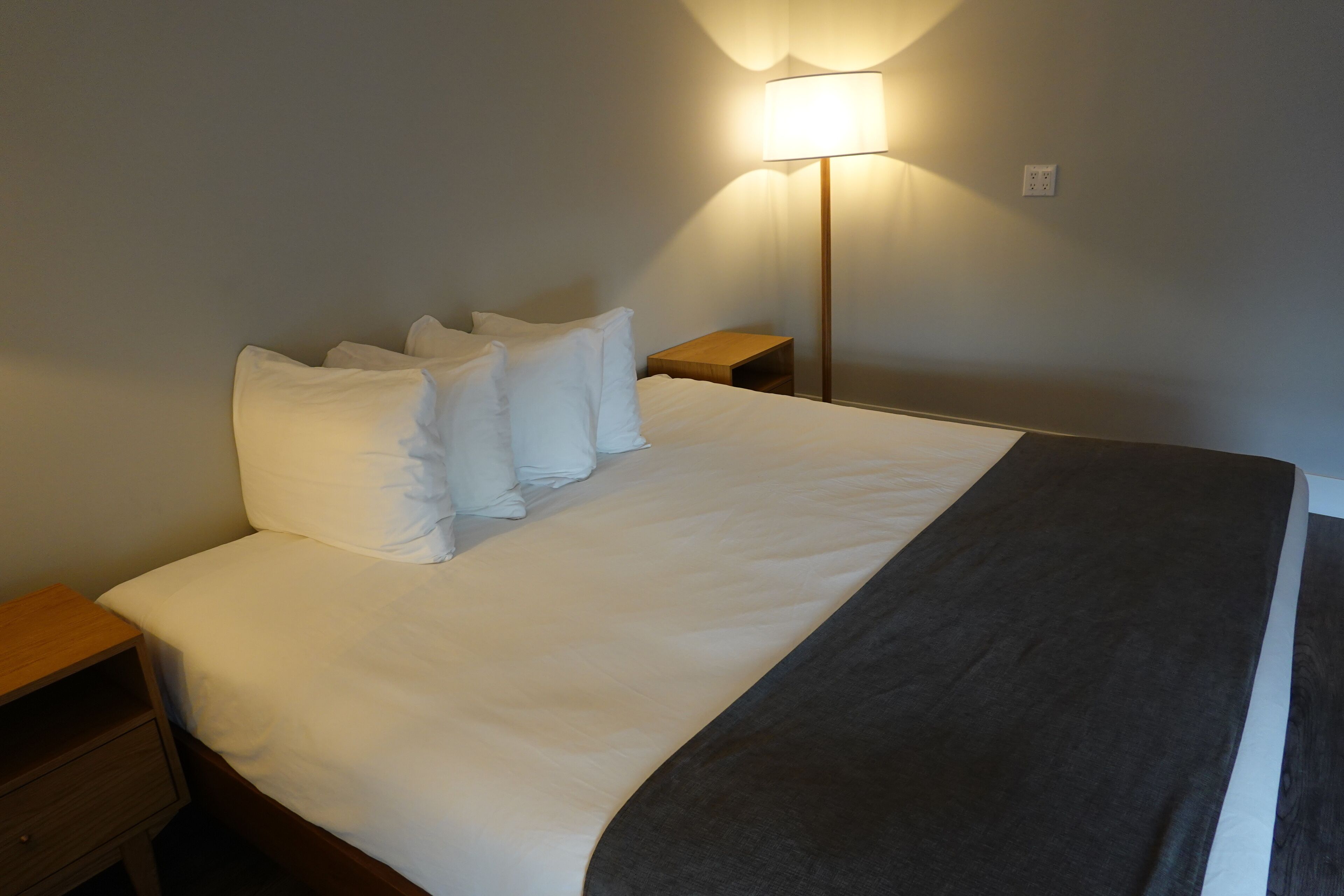 A neatly made bed with white linens and a grey throw blanket, a bedside table with a lamp, and a wall socket.