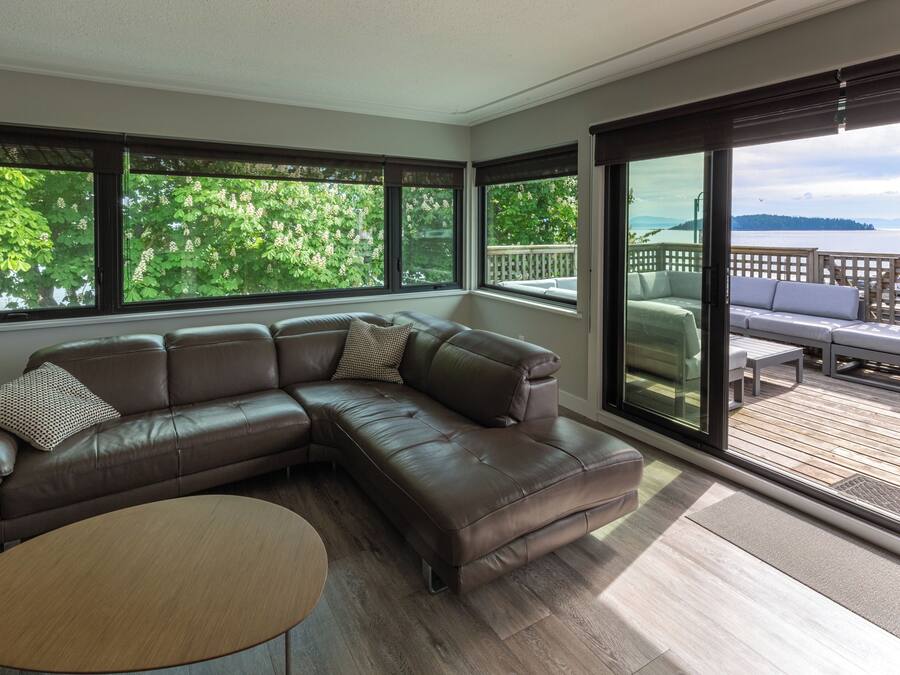 Una sala moderna con un sofá seccional, una mesa de centro redonda y ventanas amplias con vistas a una terraza y follaje.