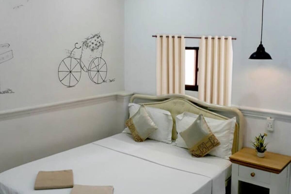 Quarto com uma cama de solteiro, uma pequena mesa de cabeceira com abajur, uma janela com cortinas e uma bicicleta desenhada na parede.