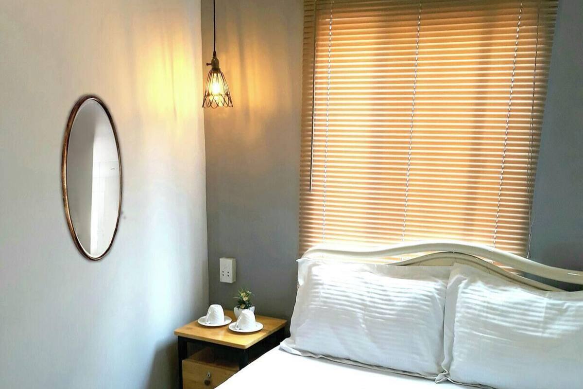 Uma cama bem arrumada com lençóis brancos, uma mesinha de cabeceira com abajur e uma janela com persianas.