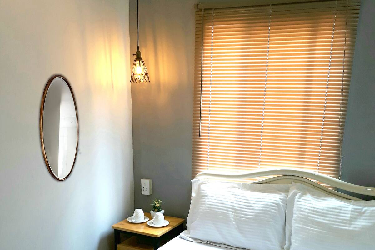 Uma cama de casal com roupa de cama branca, uma mesa de cabeceira com abajur e uma janela com persianas.