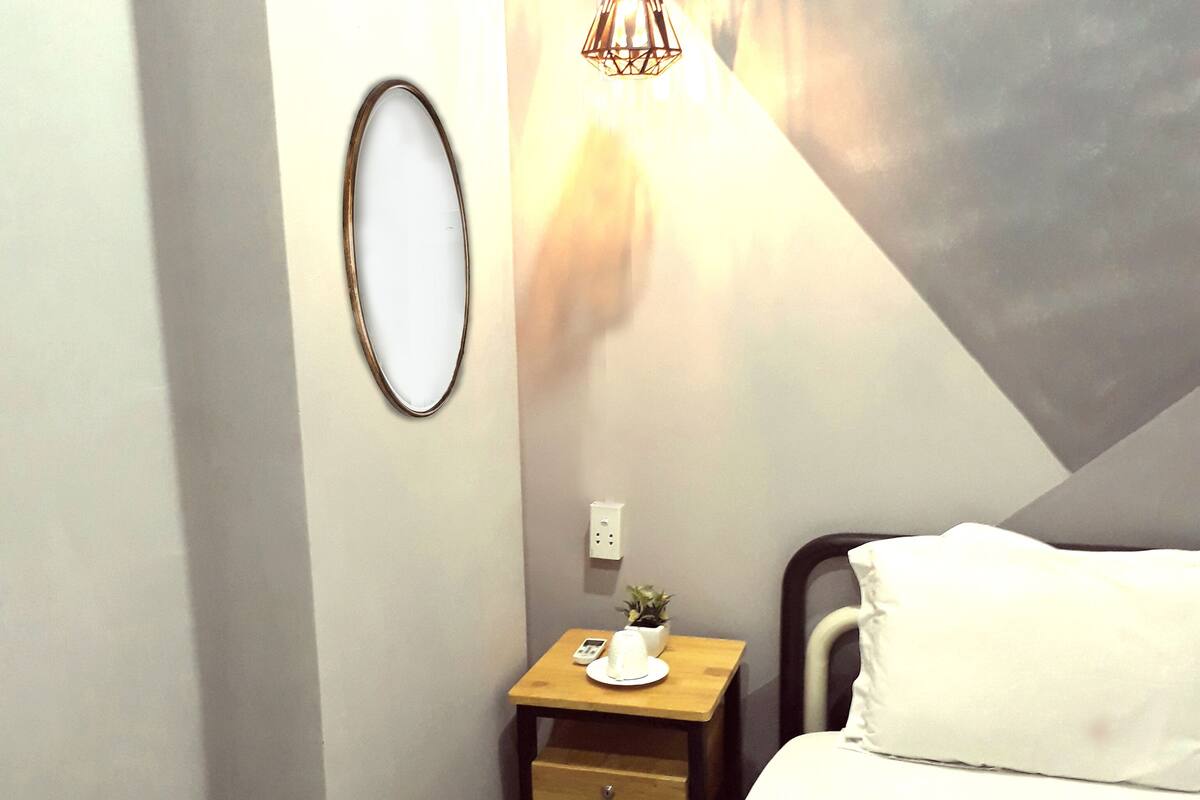 Um quarto individual com cama, mesa de cabeceira, luminária, uma pequena planta e espelho fixado na parede.
