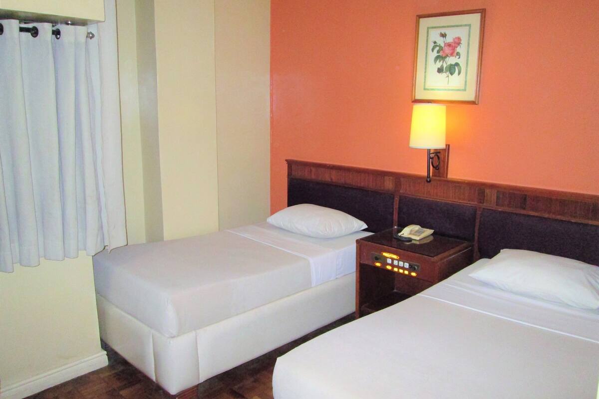 A hotel room with two single beds, a nightstand with a phone, a wall-mounted lamp, and a framed floral picture.