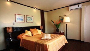 A hotel room with a bed, two bedside tables, a nightstand, a lamp, a vase with flowers, and a wall-mounted air conditioner. - Manila Manor Hotel (Manila)