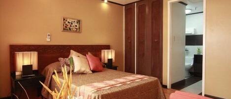 Habitación de hotel con cama, mesitas de noche, espejo, puerta y vista al baño.