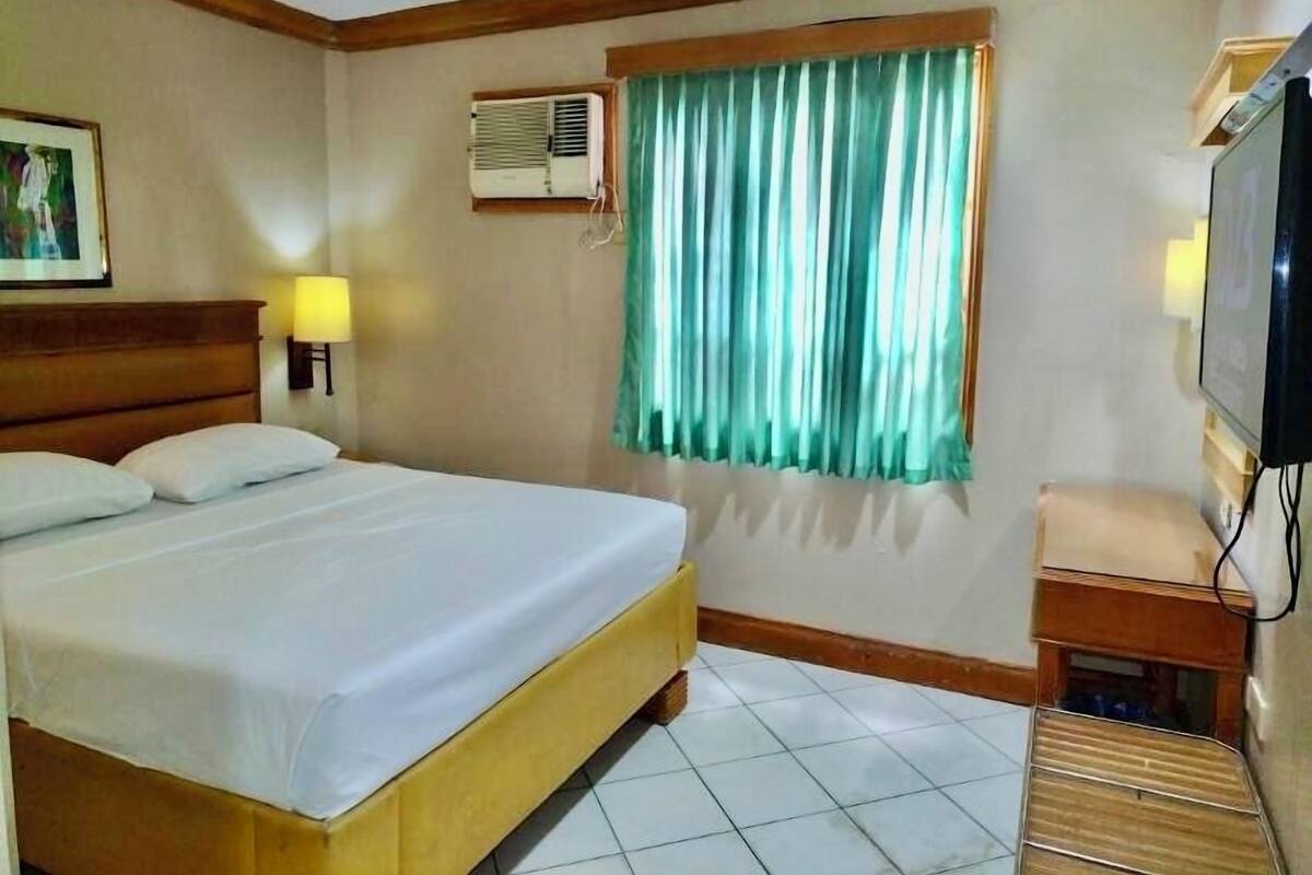 Ein Hotelzimmer mit Bett, einem fenster mit Vorhängen, einer Klimaanlage, einem Fernseher und einem kleinen Tisch.