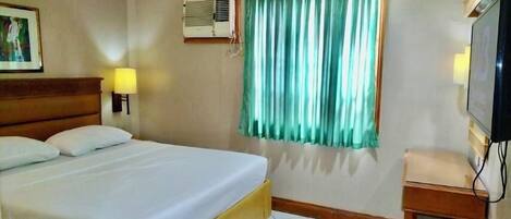 Una habitación de hotel con cama, ventana con cortinas, aire acondicionado, televisión y una mesita.