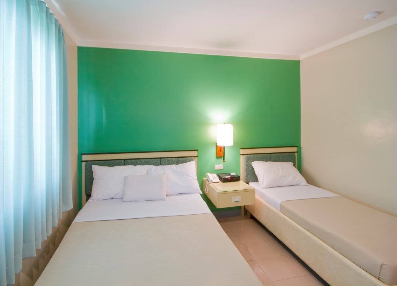 Una habitación de hotel con dos camas, una mesita de noche con lámpara y una pared verde.