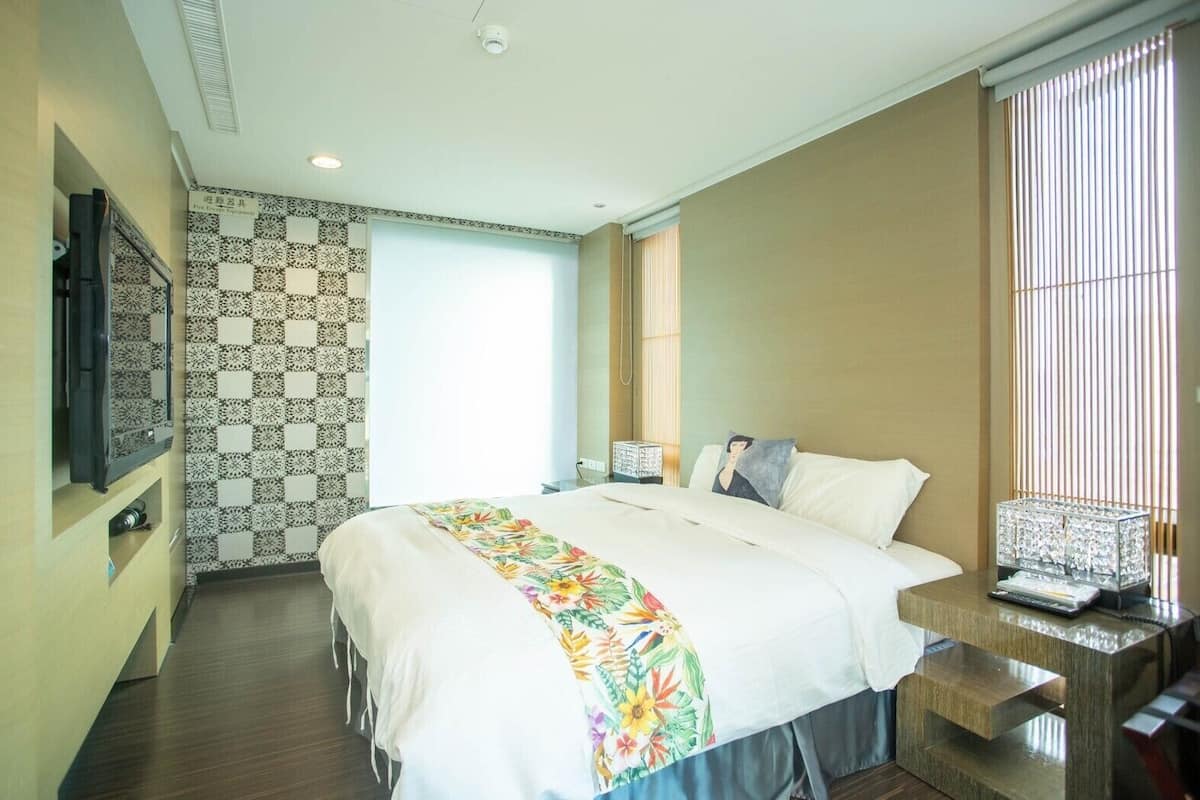 Quarto de hotel com uma cama grande, televisão na parede, mesa de cabeceira com abajur e janela com persianas.
