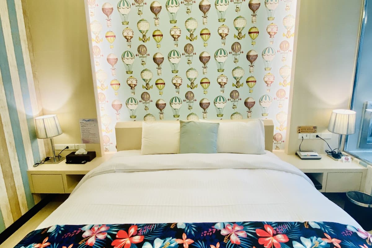 Quarto de hotel com uma cama grande, cabeceira com desenho de balão de ar quente, colcha florida e mesa de cabeceira com luminária.
