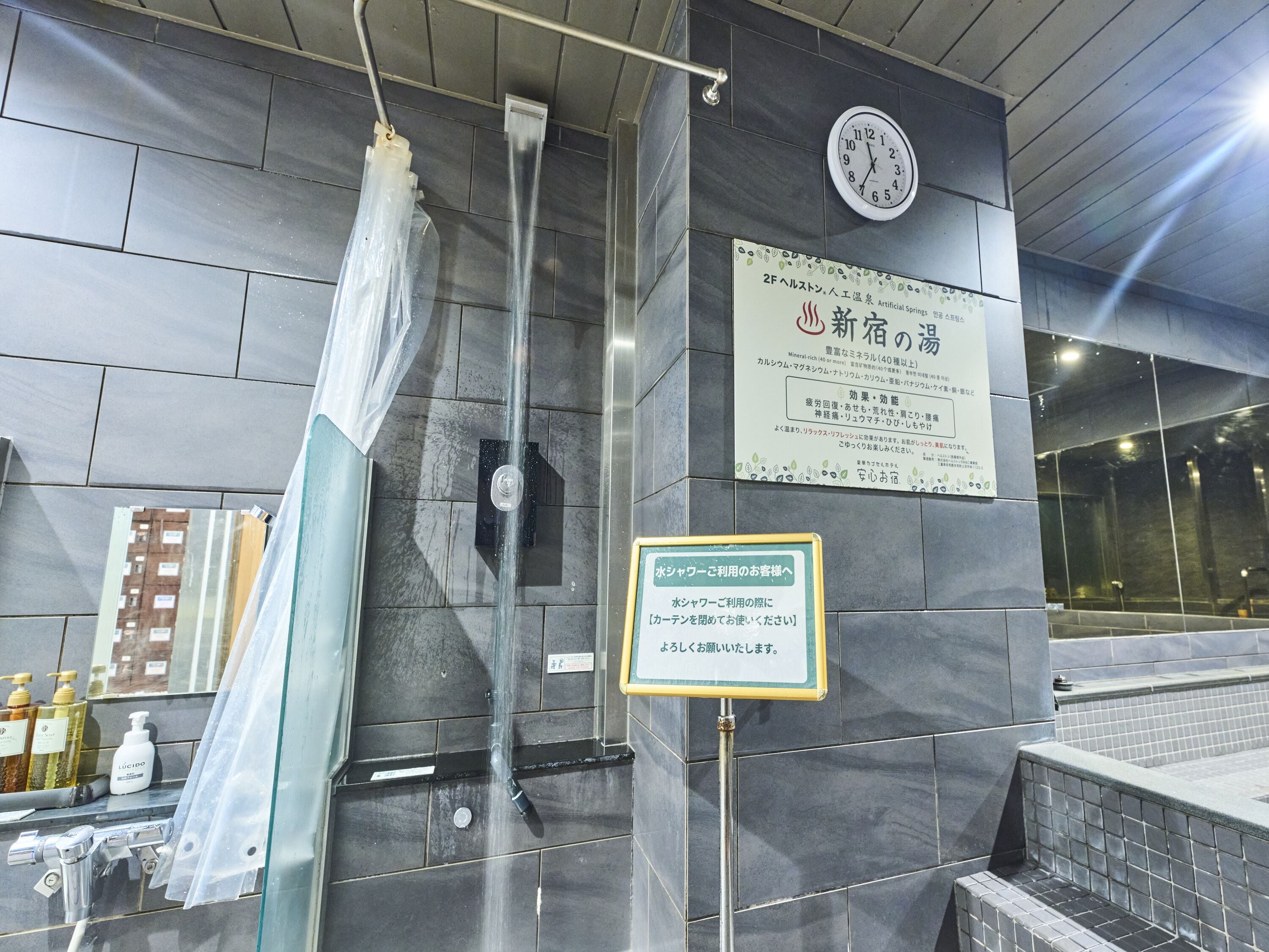 A modern bathroom with a glass shower enclosure, a clock, and informational signs in Japanese.