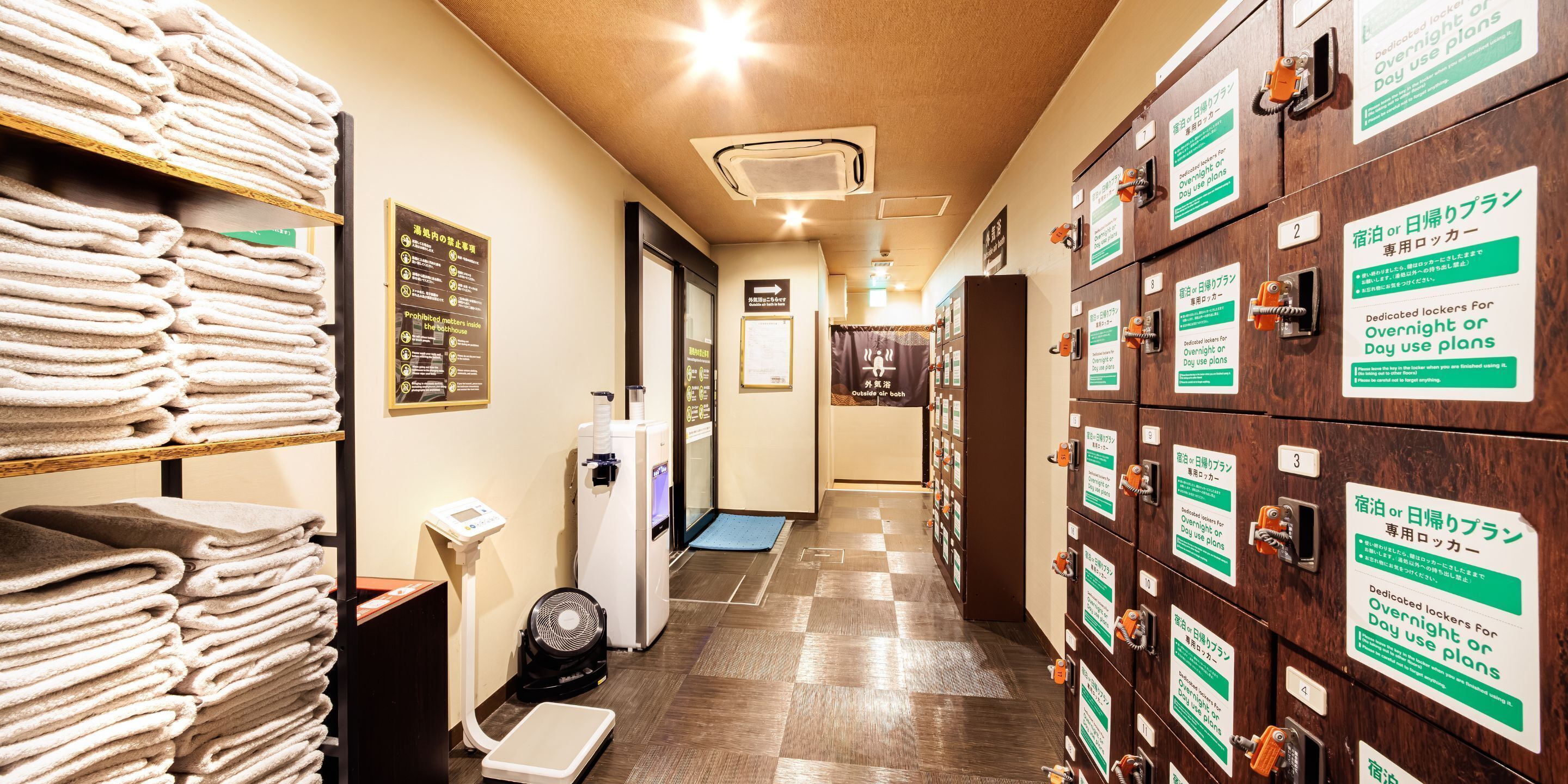 A hotel hallway with stacks of towels, a scale, a fan, and lockers with signs for overnight or day use plans.