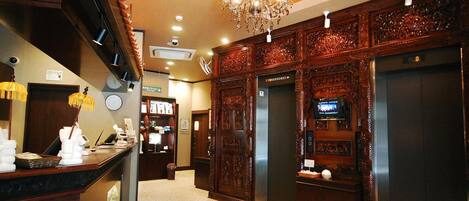 Vestíbulo de hotel con una recepción, una araña de cristal, un ascensor y paneles de madera decorativos.