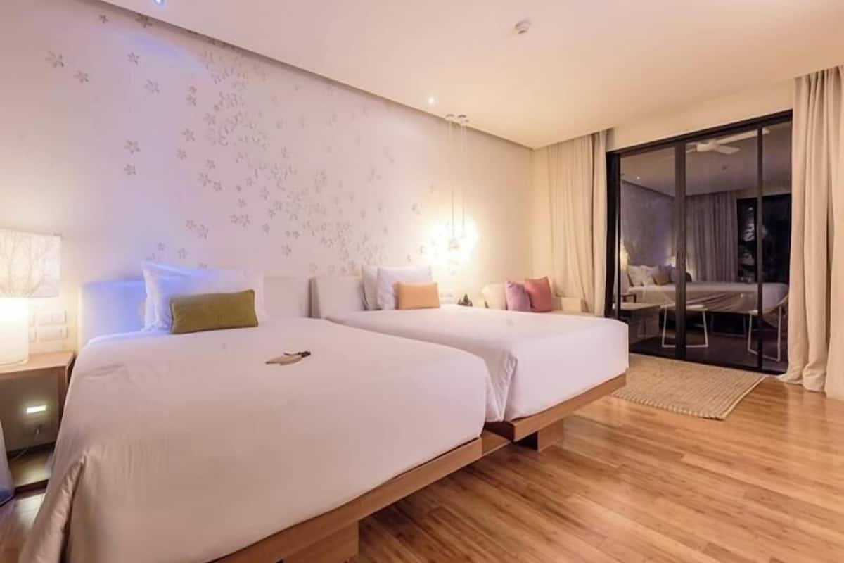 Habitación de hotel con dos camas, piso de madera, un ventanal grande con cortinas y un puertas corredera de vidrio que da a un balcón.