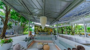 Un área de estar al aire libre y cubierta, con diseño moderno, que incluye un banco, cojines y elementos decorativos como una lámpara colgante y plantas en macetas.