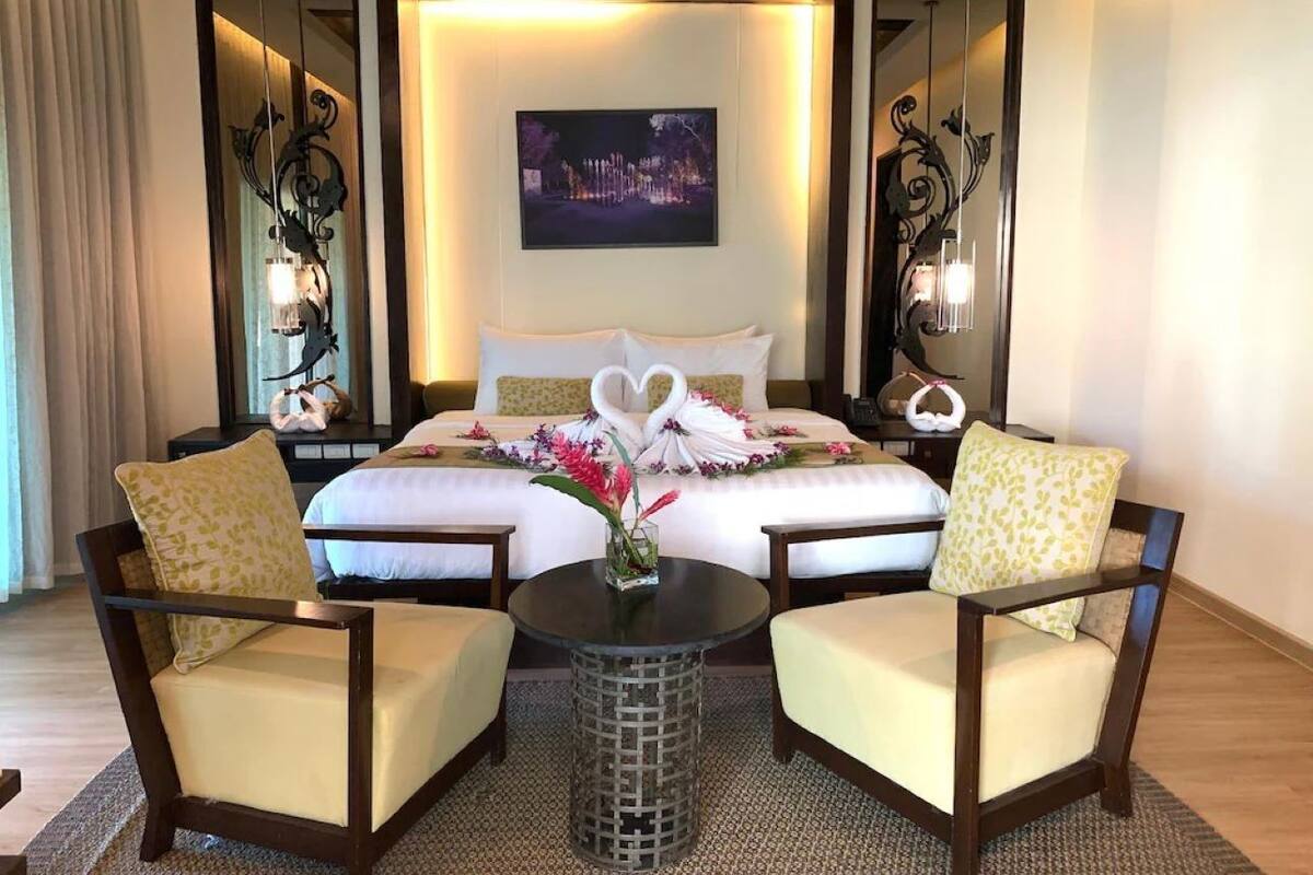 Habitación de hotel con una cama grande, dos sillones, una mesita de noche con un jarrón de flores y un cuadro colgado en la pared.