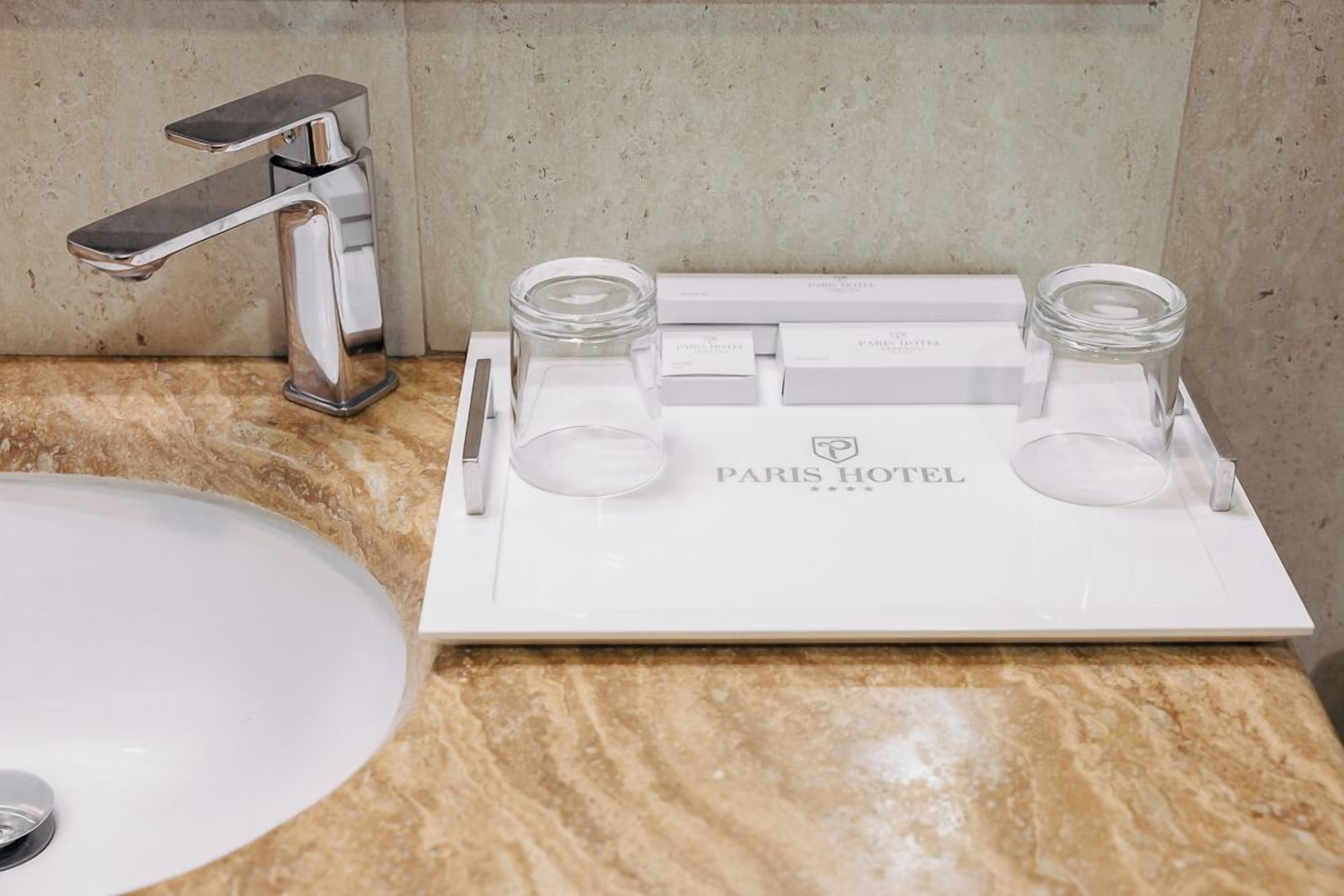 Un baño con un lavamanos blanco, un grifo cromado y una bandeja con dos frascos de vidrio y un bolígrafo. La bandeja tiene el texto 'PARIS HOTEL'.