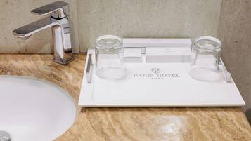 Un baño con un lavamanos blanco, un grifo cromado y una bandeja con dos frascos de vidrio y un bolígrafo. La bandeja tiene el texto 'PARIS HOTEL'.