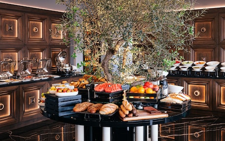 A buffet with various food items, including fruits, bread, and pastries, arranged on a black table with a large tree in the center.
