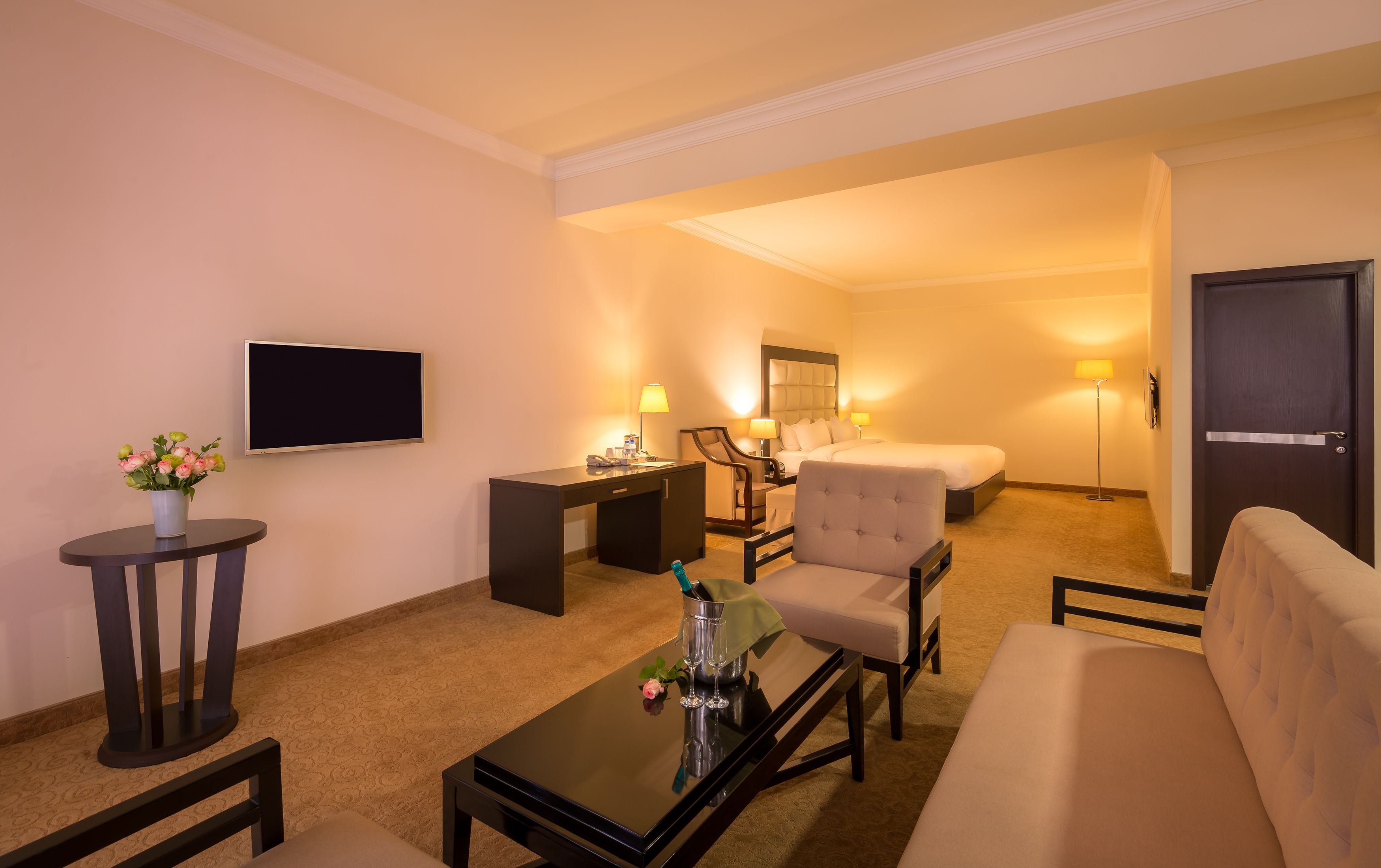 A hotel room with a flat-screen TV, a desk with a chair, a sofa, a bed, and a bedside table with a lamp.