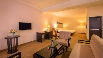 A hotel room with a flat-screen TV, a desk with a chair, a sofa, a bed, and a bedside table with a lamp.