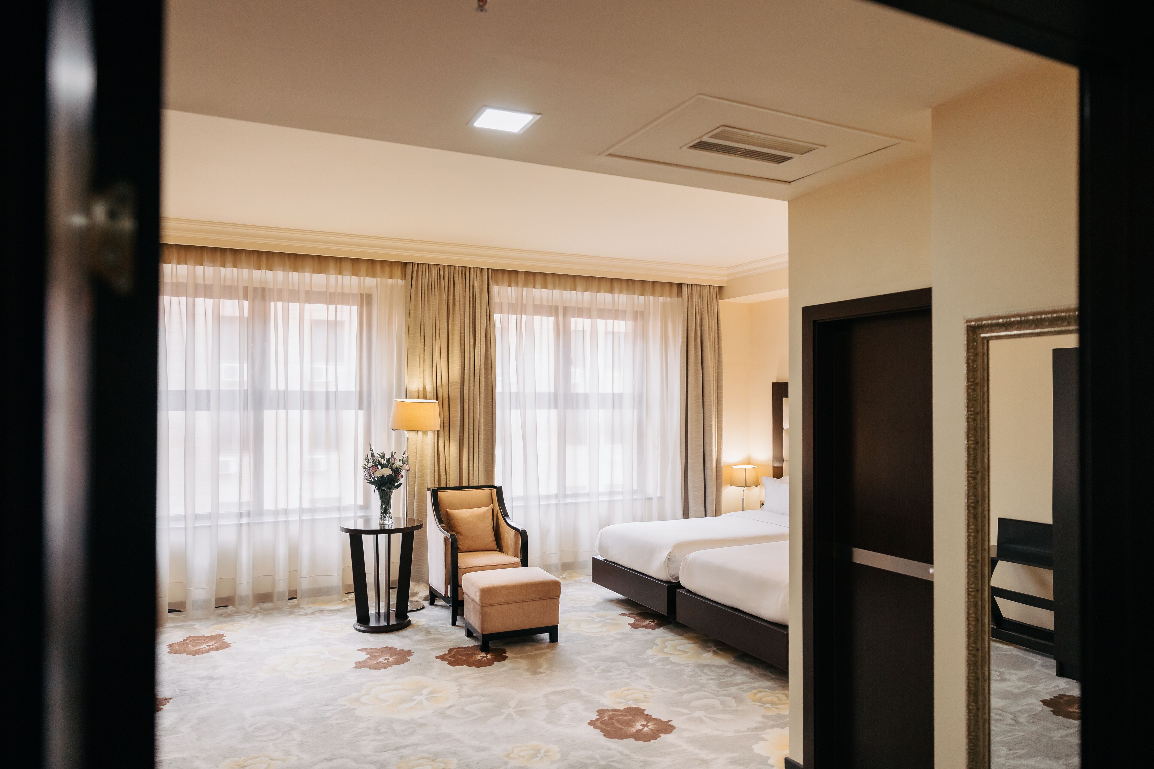 Une chambre d’hôtel avec un lit, une chaise, une petite table avec une lampe et une grande fenêtre avec des rideaux.