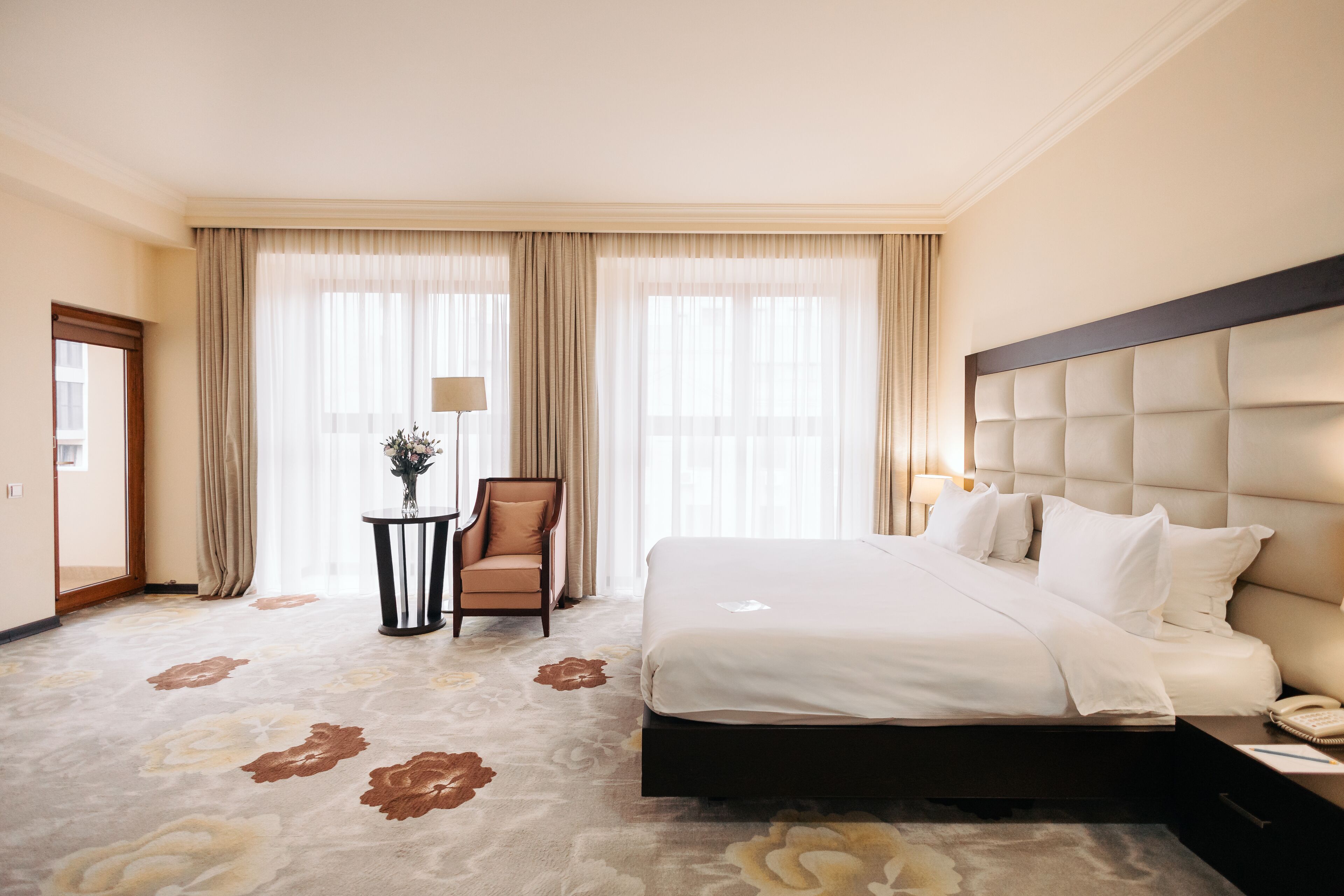 Habitación de hotel con una cama grande, una silla, una mesita con un florero y cortinas en las ventanas.