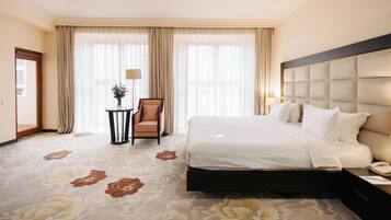 Habitación de hotel con una cama grande, una silla, una mesita con un florero y cortinas en las ventanas.