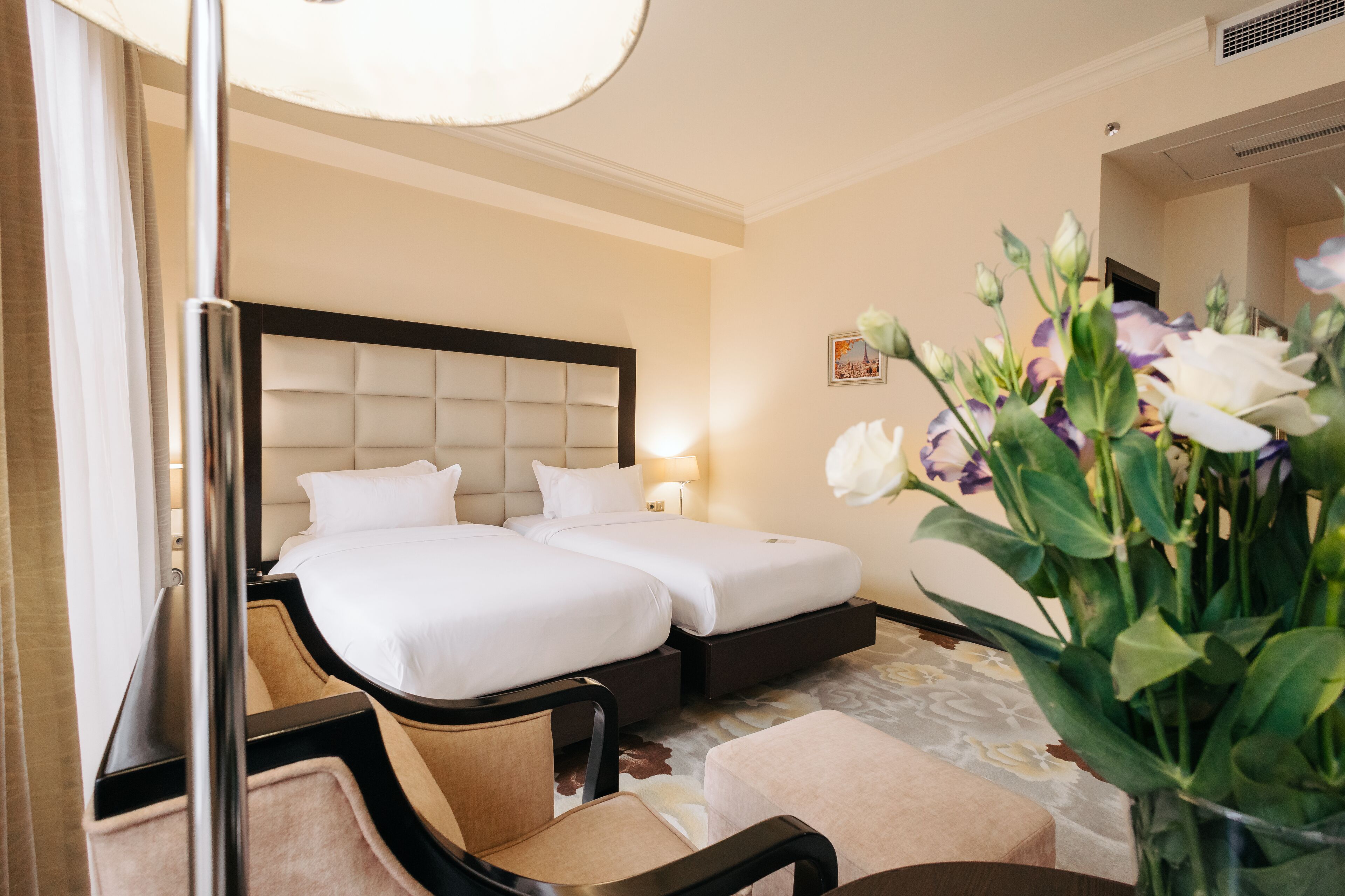 A hotel room with two beds, a sitting area with a chair, a bedside table, a lamp, and a vase with flowers.
