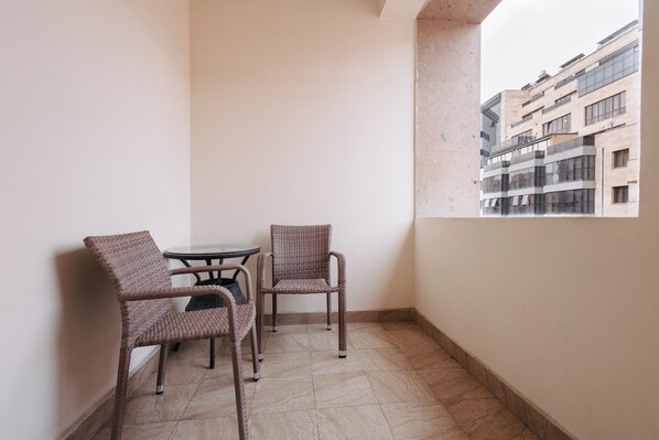 A balcony with two wicker chairs and a small round table, a tiled floor, and a view of a building through a window.