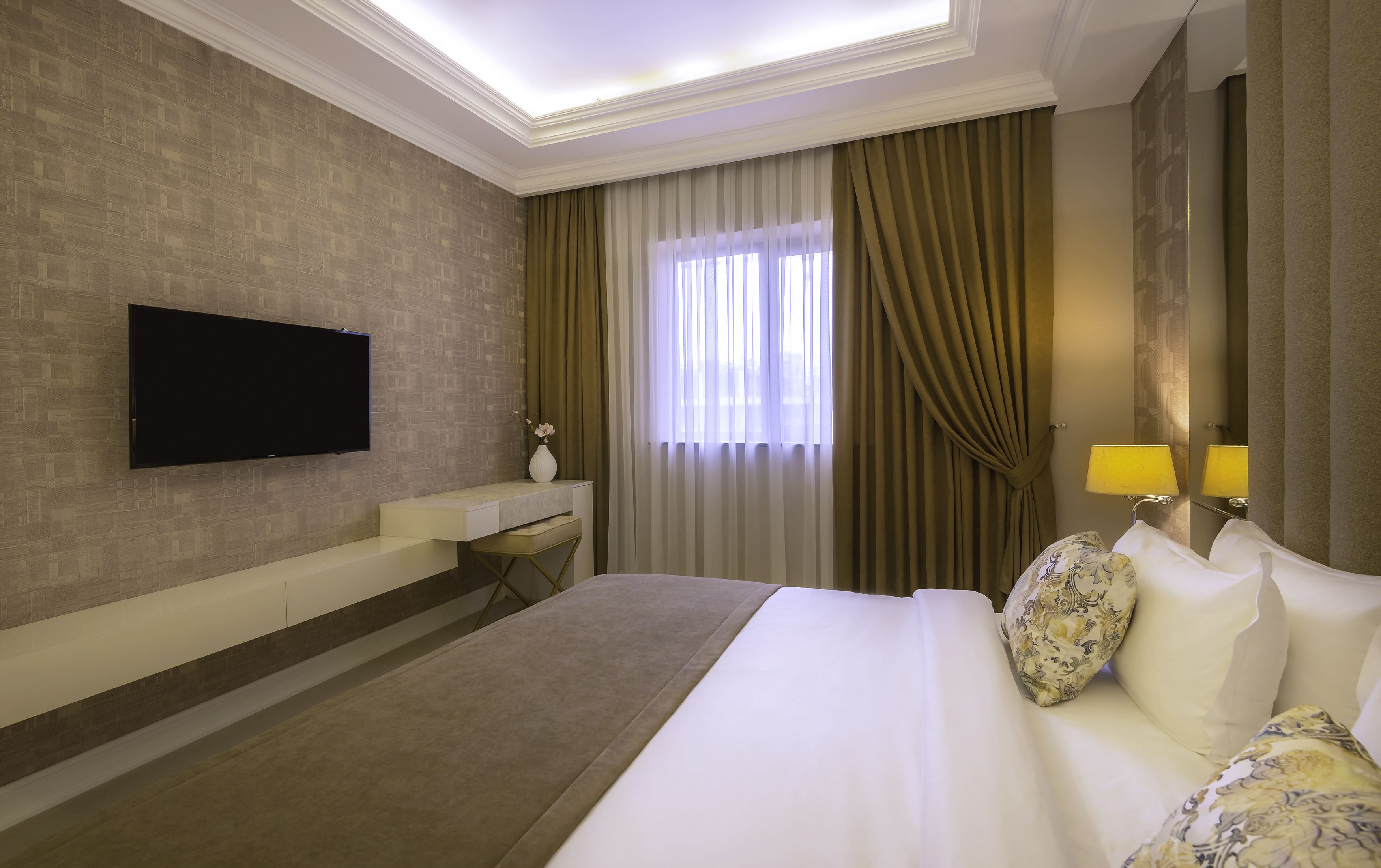 Habitación de hotel con televisión de pantalla plana en la pared, un escritorio con silla, cama con almohadas estampadas y una ventana con cortinas.